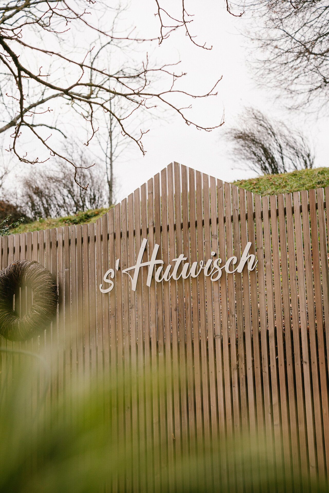 Holzzaun mit dem Schriftzug ‚s’Hutwisch‘, dahinter kahle Bäume in herbstlicher Stimmung.
