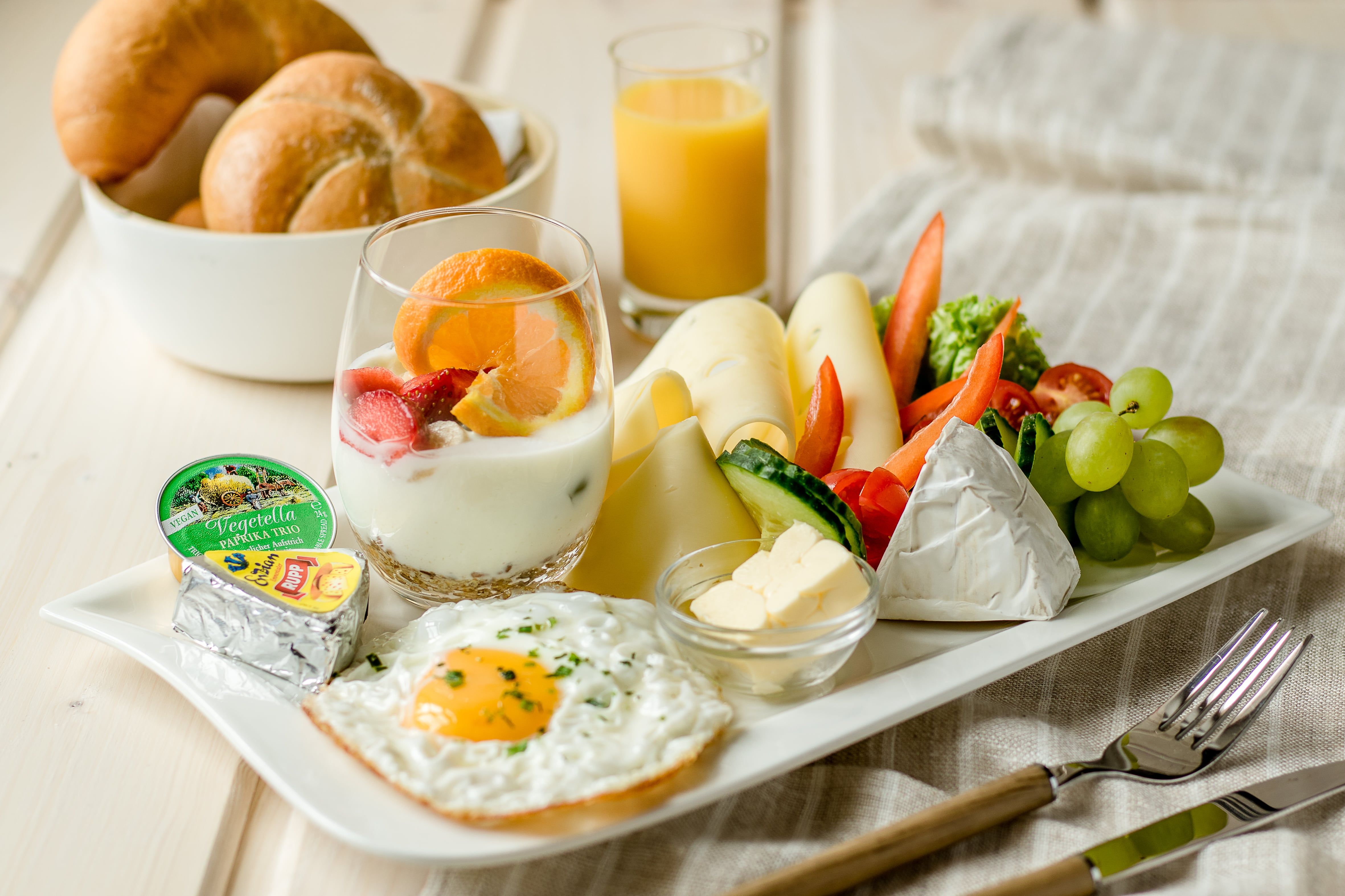 Ein Teller mit vegetarischem Frühstück: Spiegelei, Käse, Gemüse, Joghurt mit Obst, Brötchen und Orangensaft.