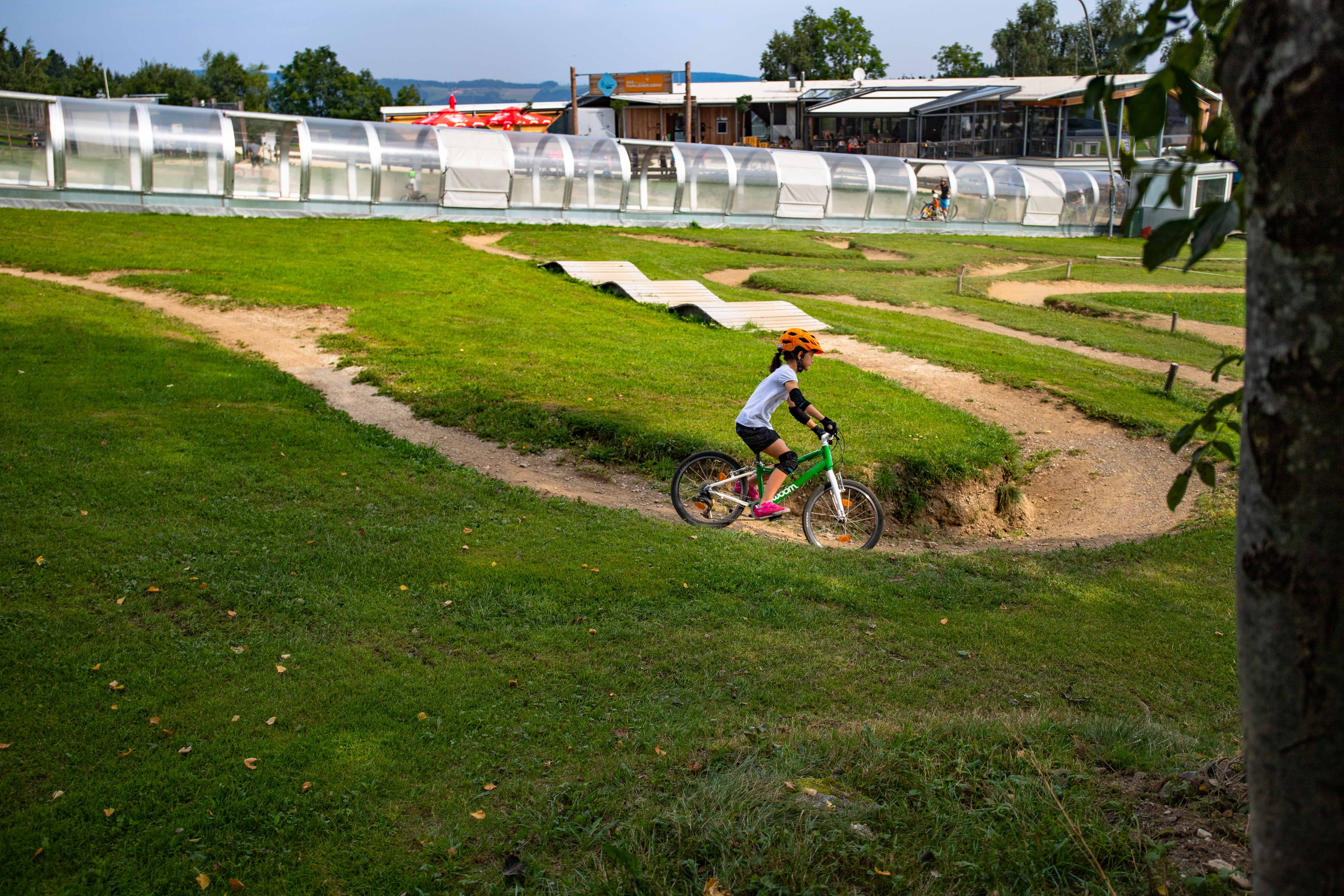 Ein Kind mit orangem Helm fährt auf einem grünen Fahrrad durch einen Mini-Bikepark mit Hügeln und Kurven.