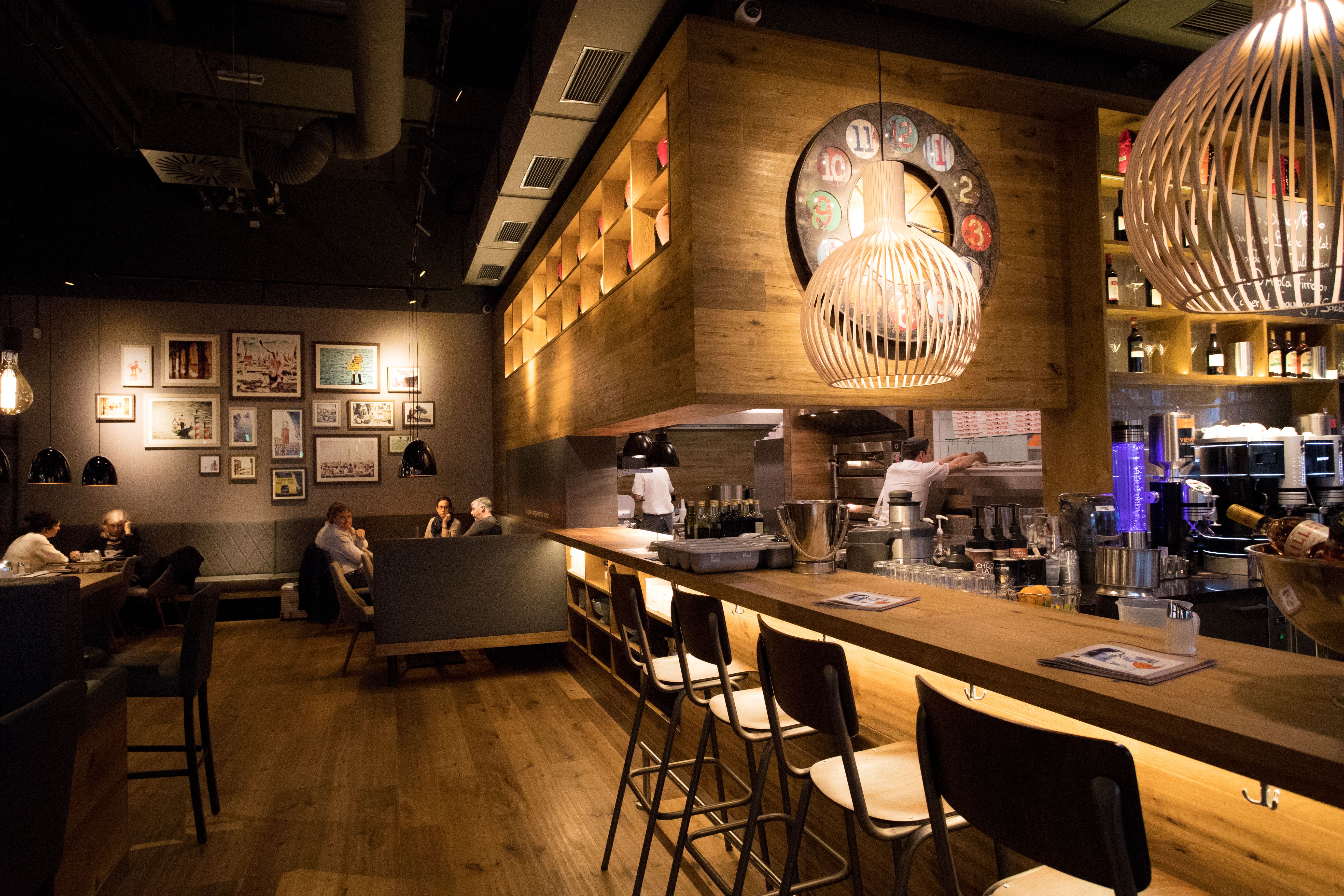 Modernes Restaurant mit Holzboden, gemütlichen grauen Sitzbänken und einer Bar aus Holz mit Barhockern.
