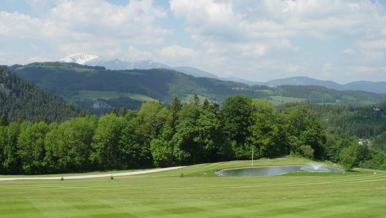 Grüne Golfplatzlandschaft mit Teich und bewaldeten Hügeln im Hintergrund.
