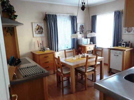 Cozy kitchen with wooden furniture, dining table and chairs, two windows with curtains, and kitchen utensils on the worktop.