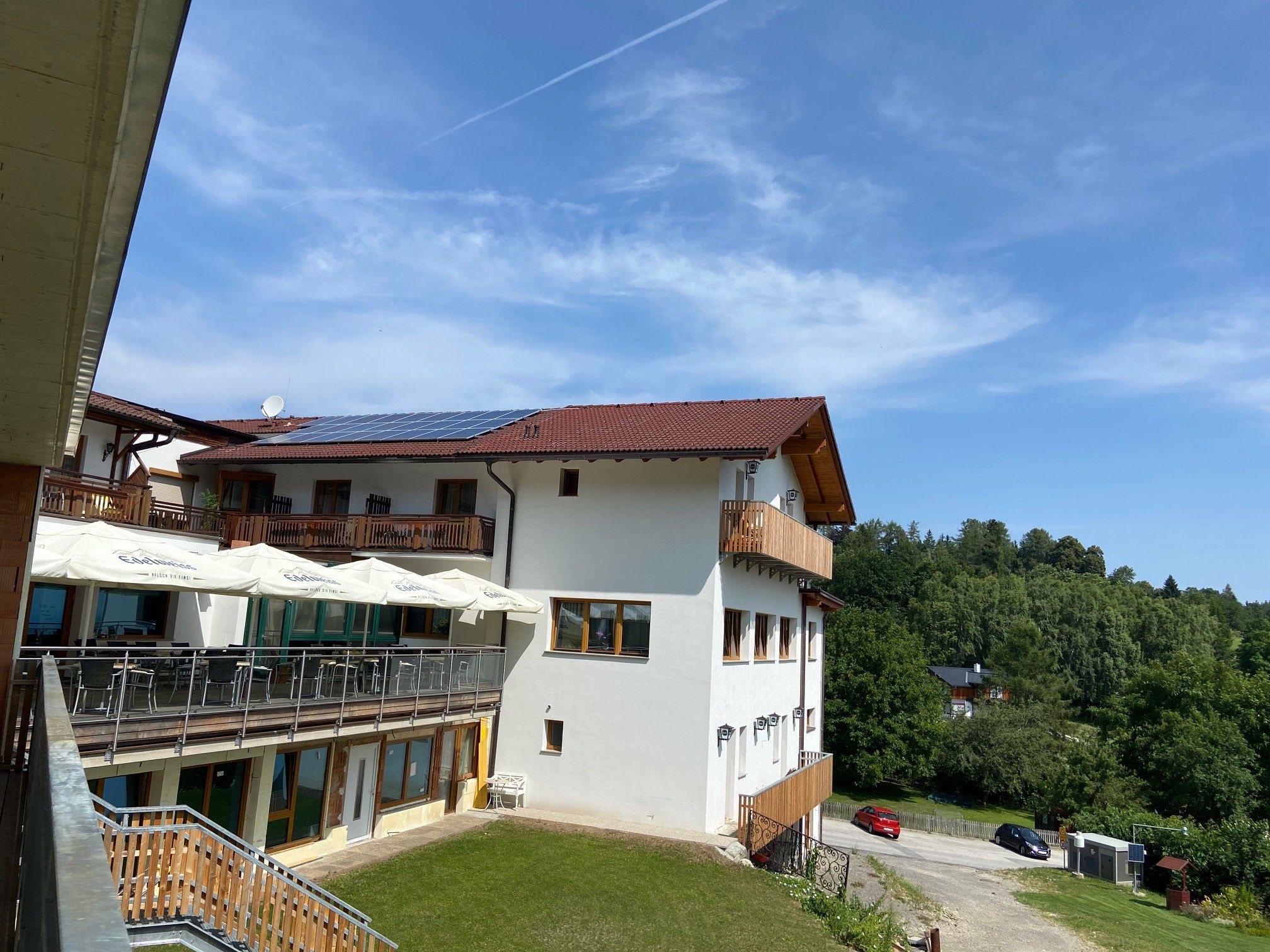 View of a building with terrace and sunshades, surrounded by green landscape and blue sky.