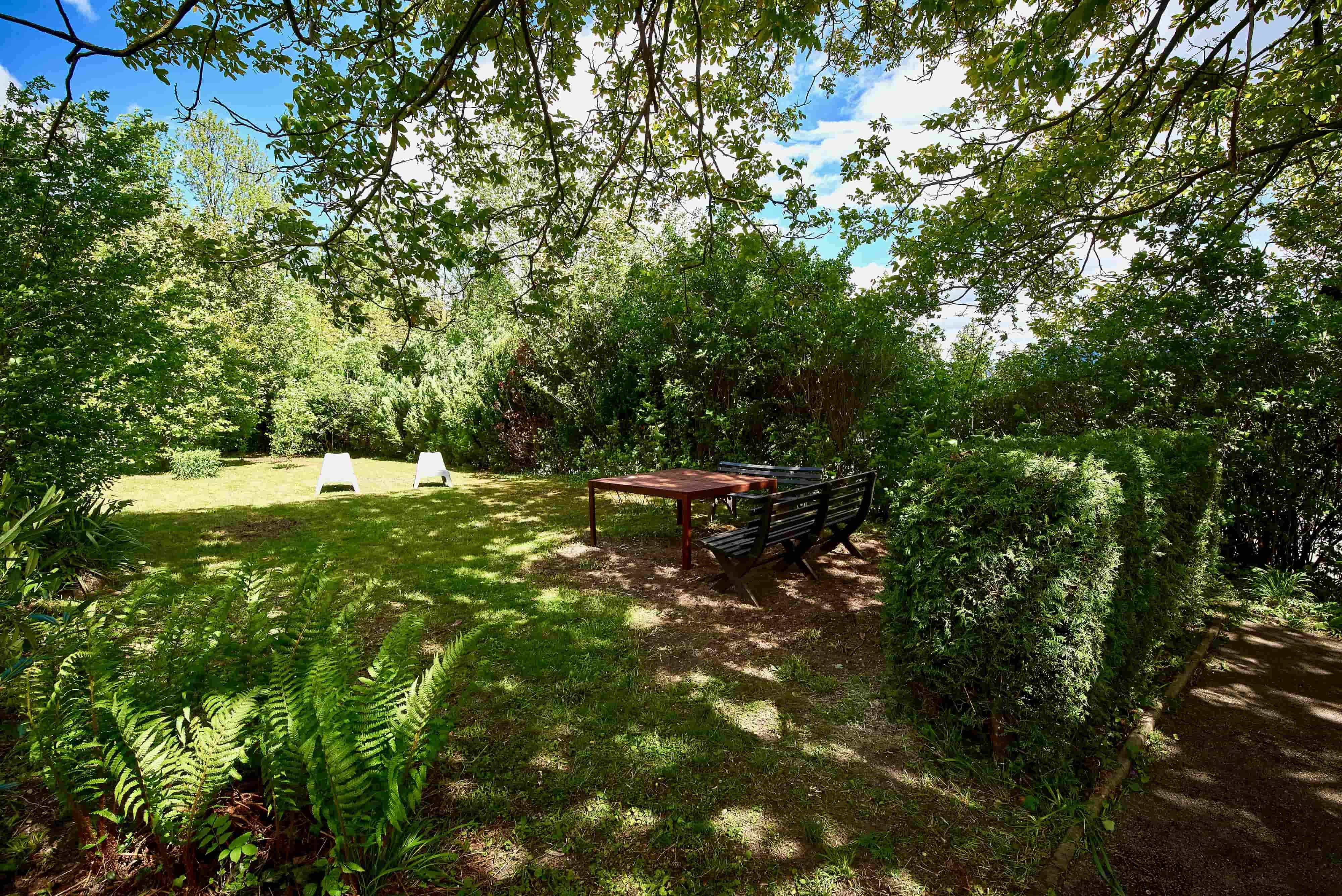 Ein grüner Garten mit Bäumen, Sträuchern, zwei weißen Liegestühlen und einer Sitzgruppe mit Tisch im Schatten.