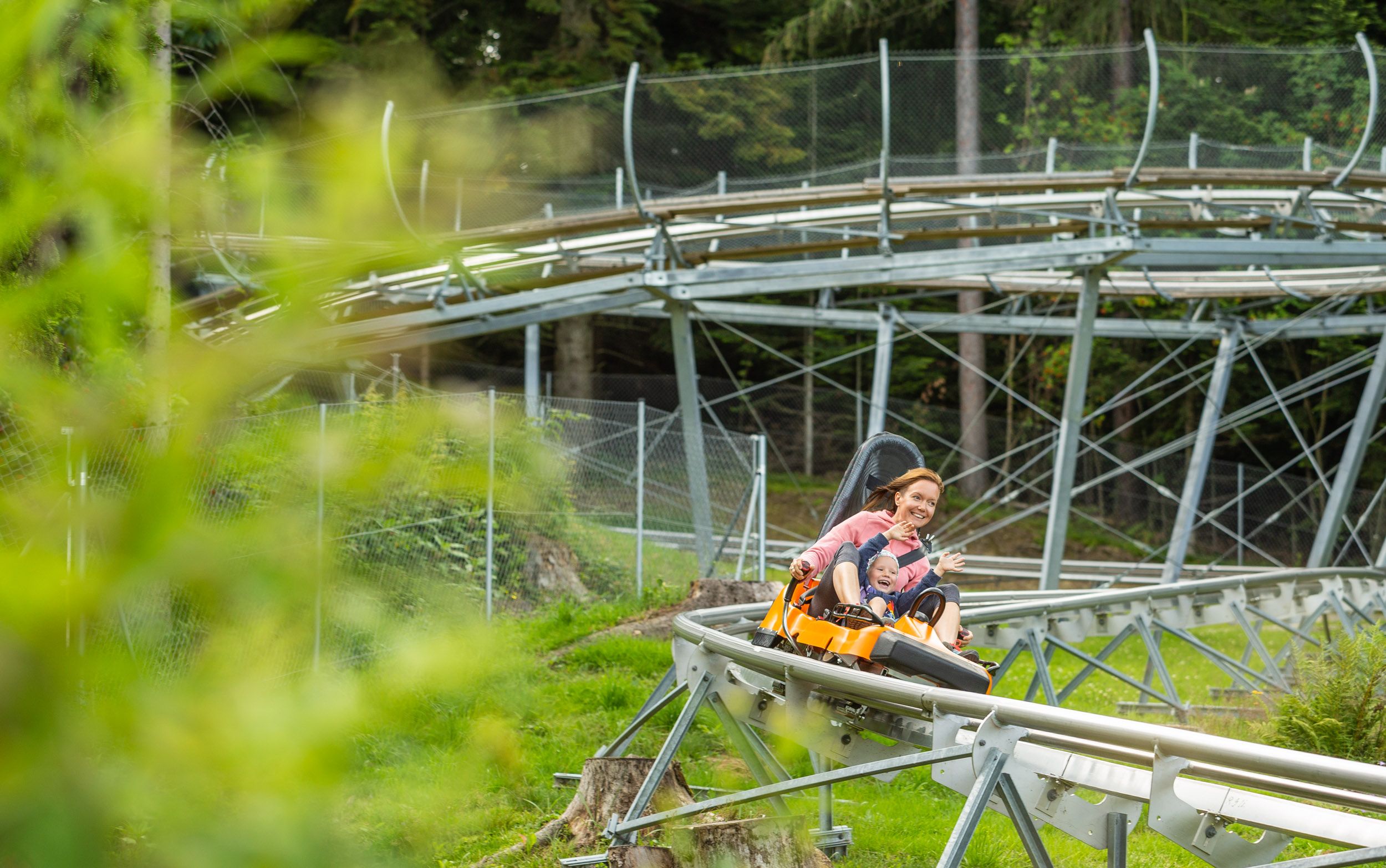 Eine Frau und ein Kind fahren lachend auf einer Sommerrodelbahn durch eine grüne Landschaft.