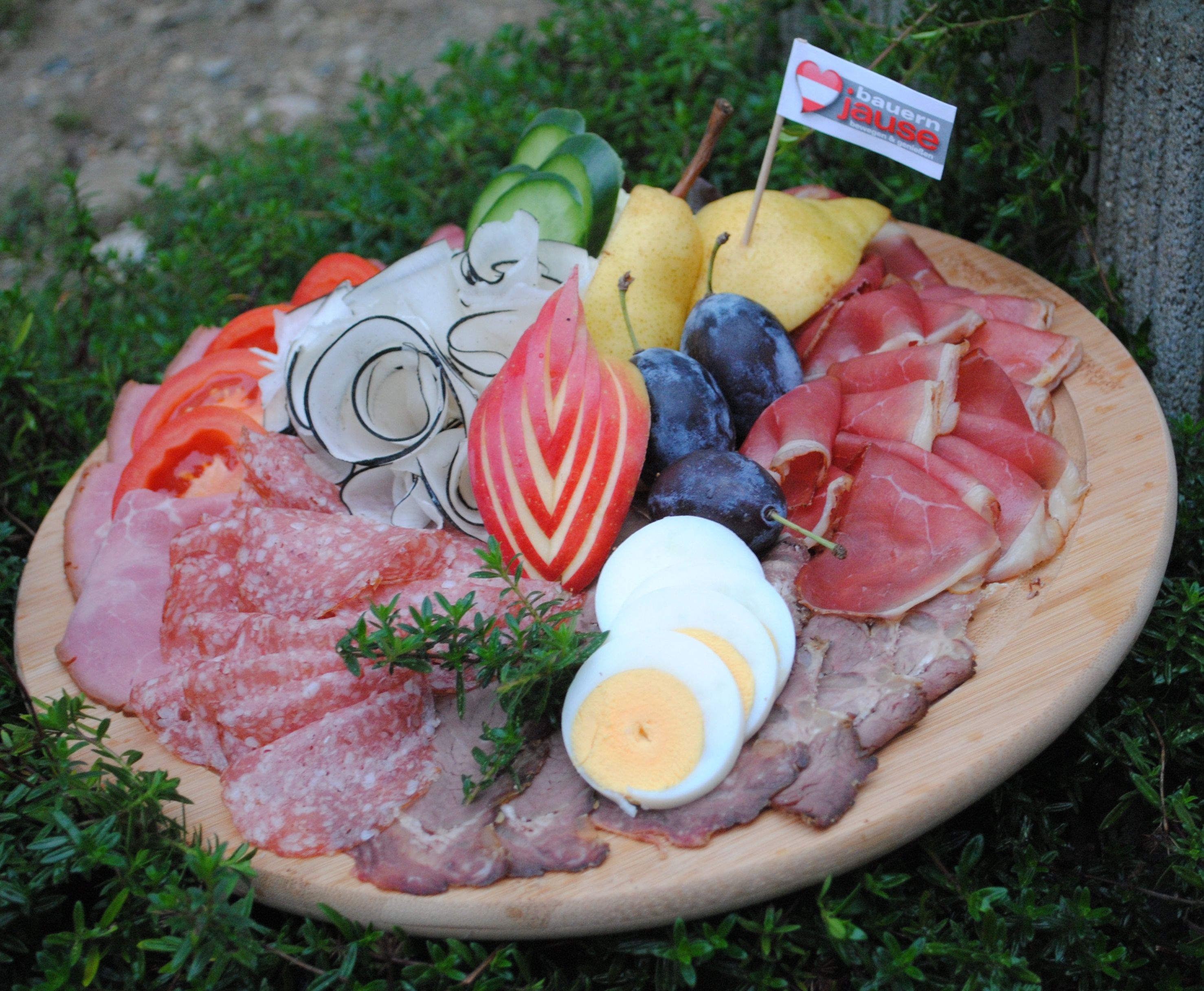 Holzplatte mit aufgeschnittenen Wurstwaren, Eiern, Gemüse und Obst auf einer grünen Wiese.