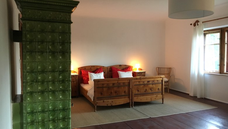 Ein gemütliches Doppelzimmer mit Holzmöbeln, einem grünen Kachelofen und einem großen Fenster mit weißen Vorhängen.