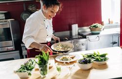 Eine Köchin hält eine Pfanne und richtet ein Teller mit einem Gericht an.