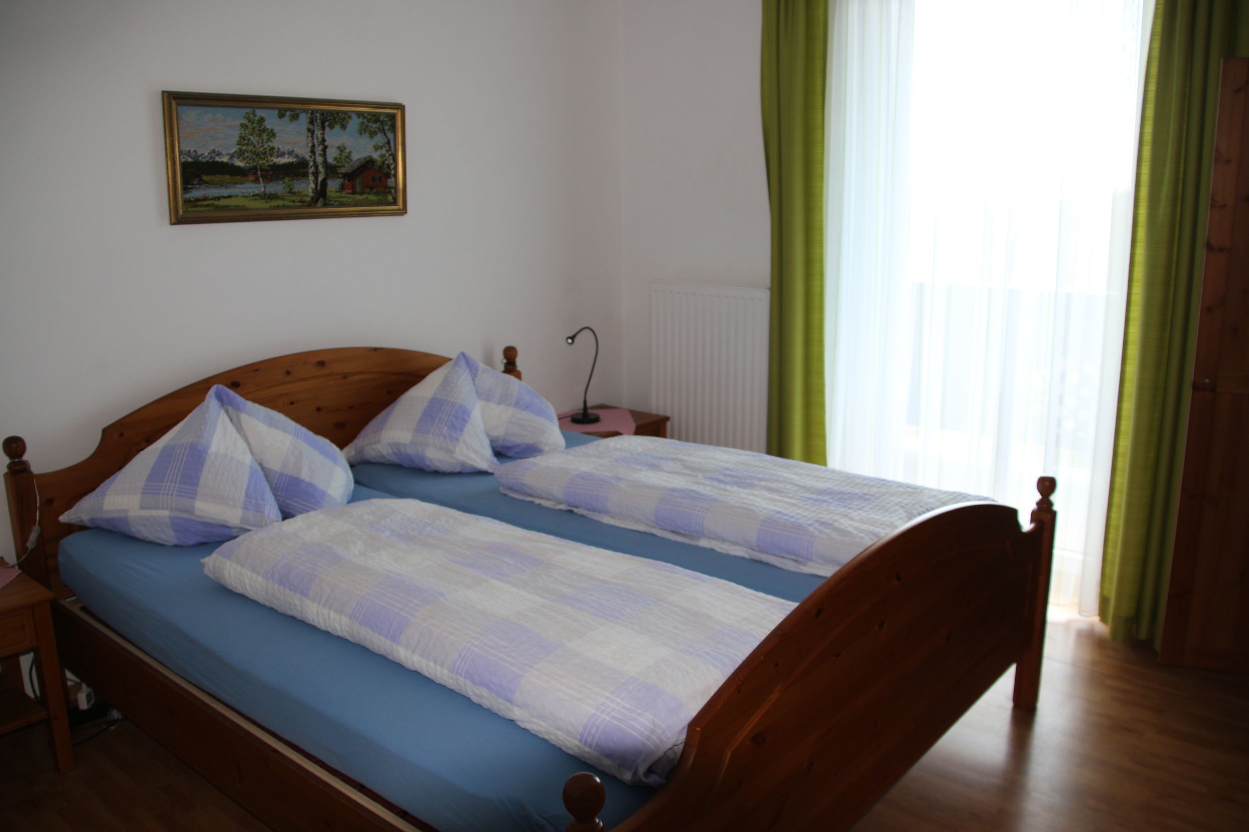 Doppelzimmer mit Holzbett, blau-weiß karierter Bettwäsche, Bild an der Wand und grünem Vorhang.