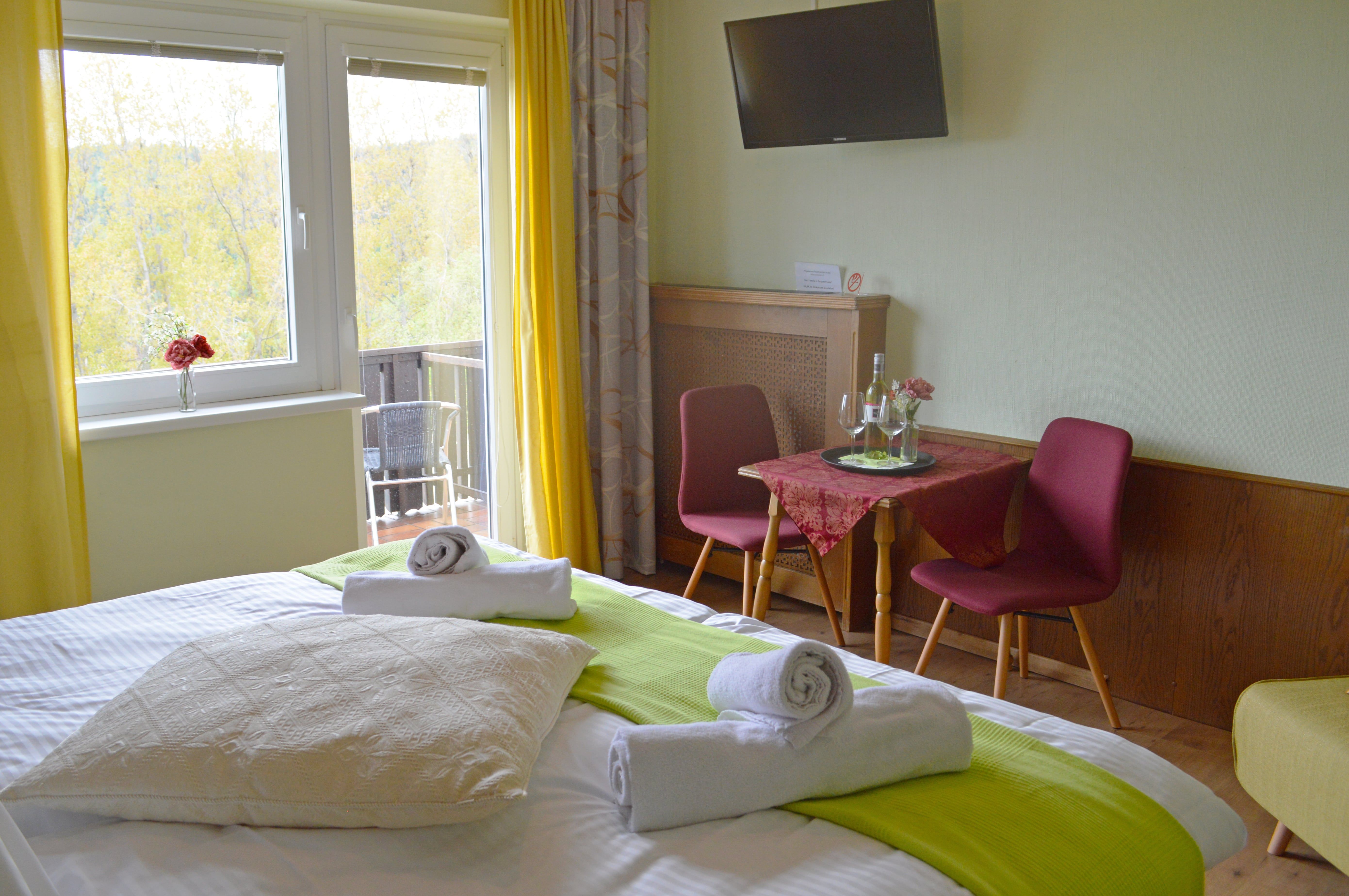 Ein gemütliches Doppelzimmer mit Bett, Tisch, Stühlen und Balkon. Gelbe Vorhänge und grüne Akzente sorgen für eine freundliche Atmosphäre.
