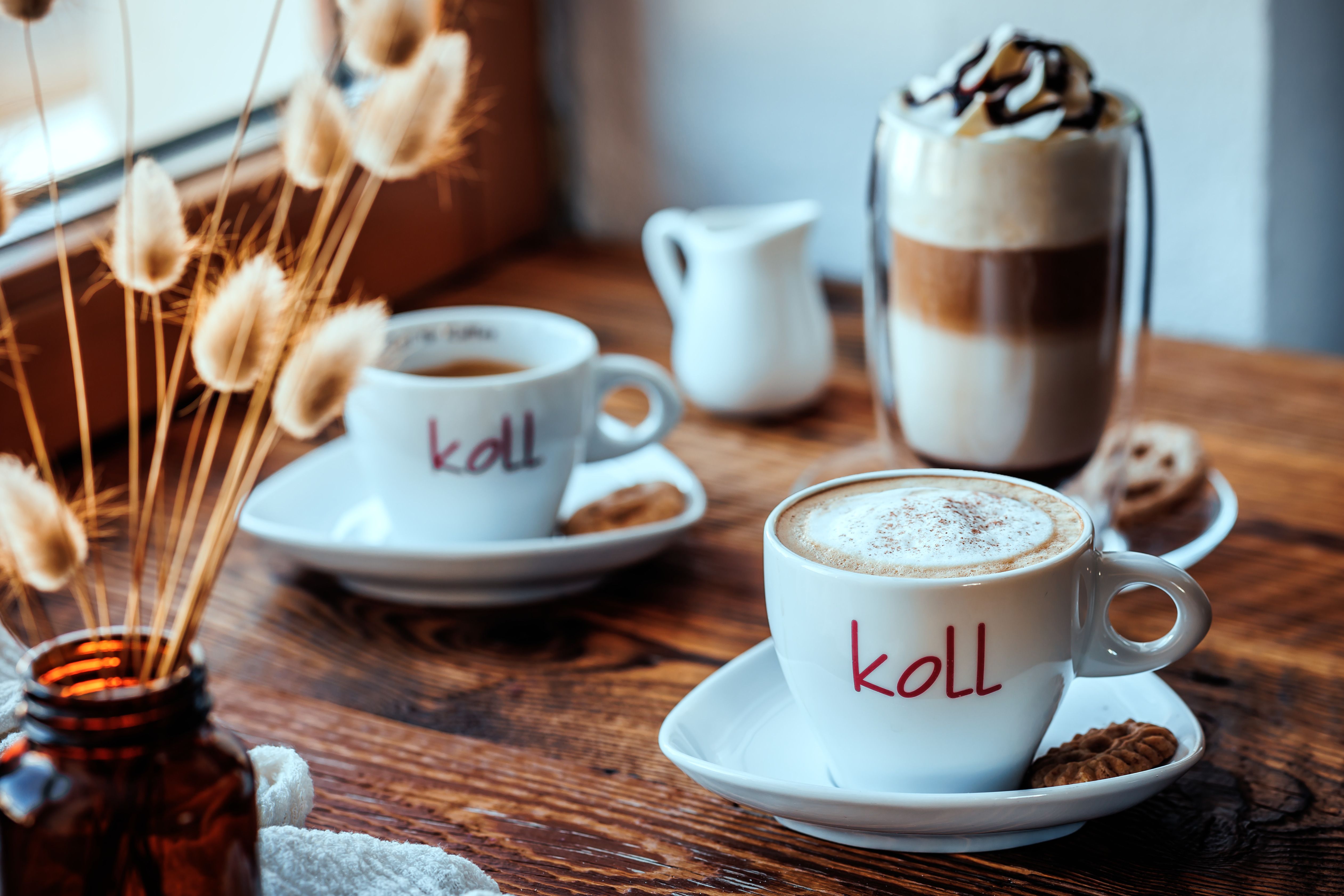 Zwei Tassen Kaffee auf einem Holztisch, eine mit Milchschaum, daneben ein Glas Latte Macchiato und eine Vase mit Trockenblumen.