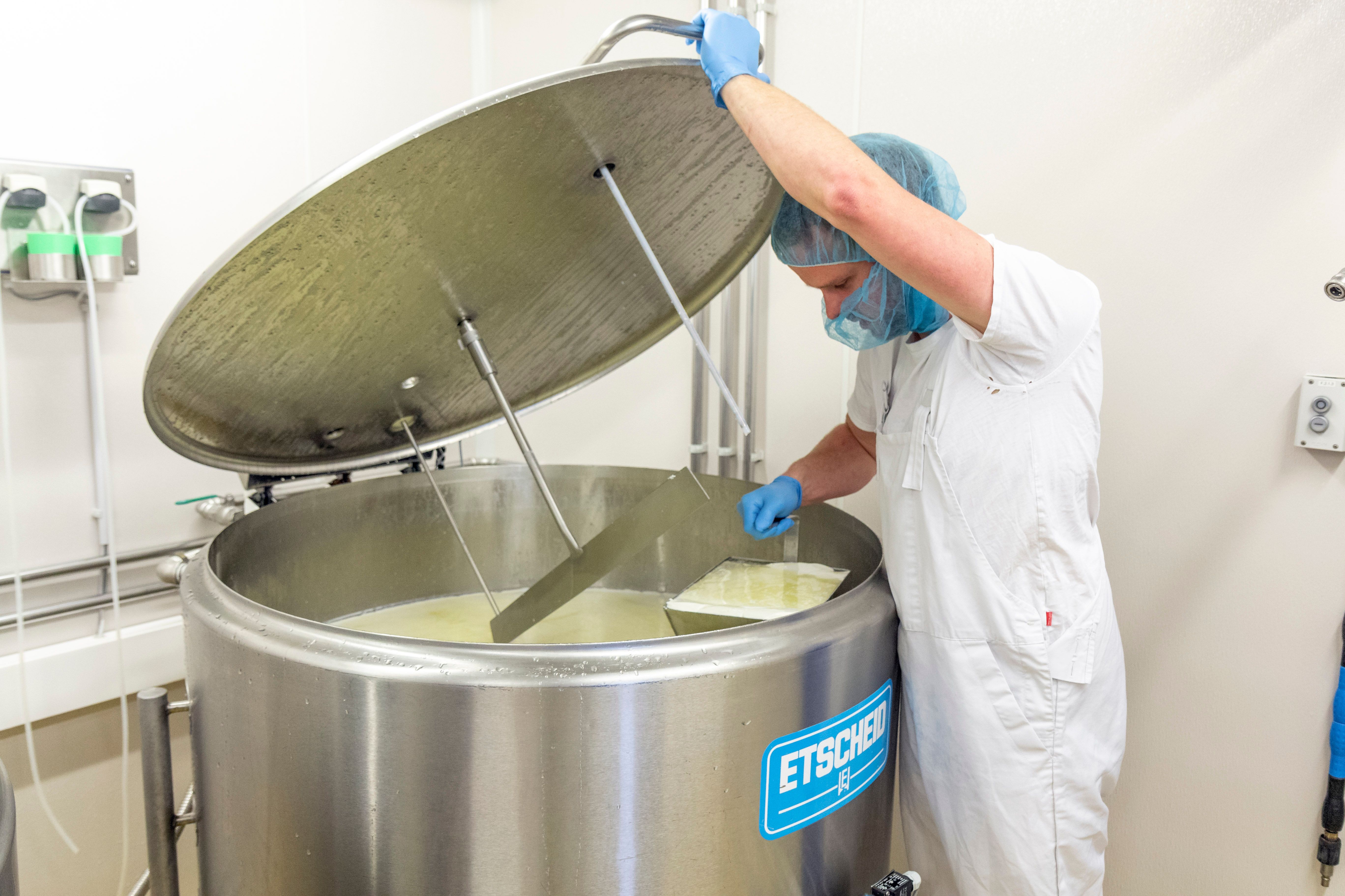 Ein Mann in Arbeitskleidung rührt Molke in einem silberenen Kessel in einer Molkerei.