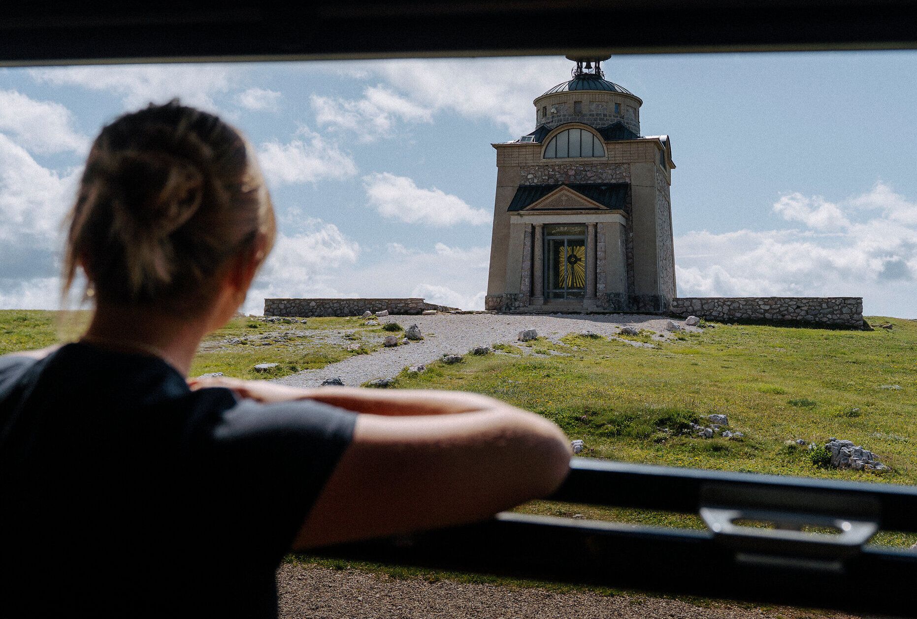Eine Frau blickt von der Schneebergbahn auf das Elisabethkircherl am Hochschneeberg. Ein Kiesweg führt zur Kirche, die umgeben von einer grünen Wiese ist, was eine Atmosphäre der Erkundung und historischer Neugier erzeugt.
