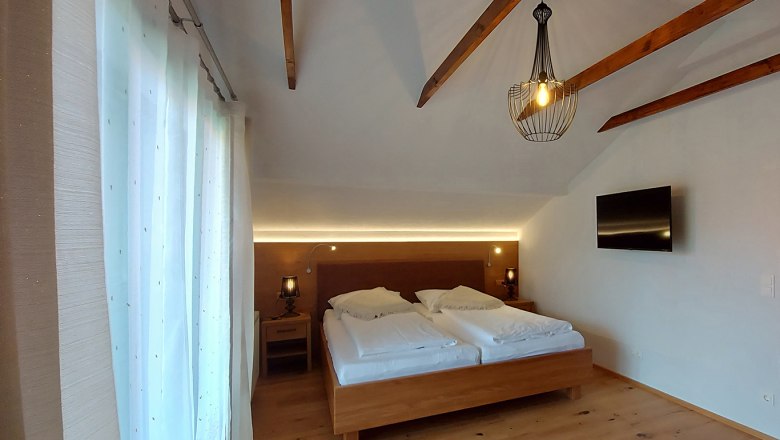 Doppelzimmer mit Holzbalken, Doppelbett, Nachttischen, Lampe und Fernseher an der Wand.