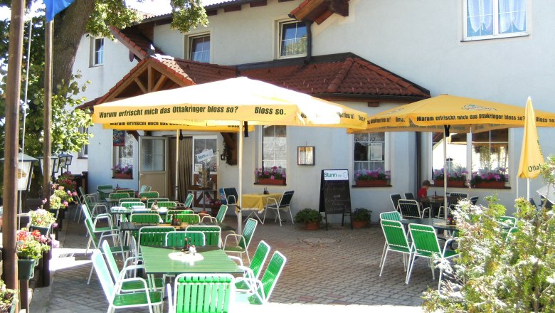 Ein Gastgarten mit grünen Stühlen und gelben Sonnenschirmen vor einem Gebäude.