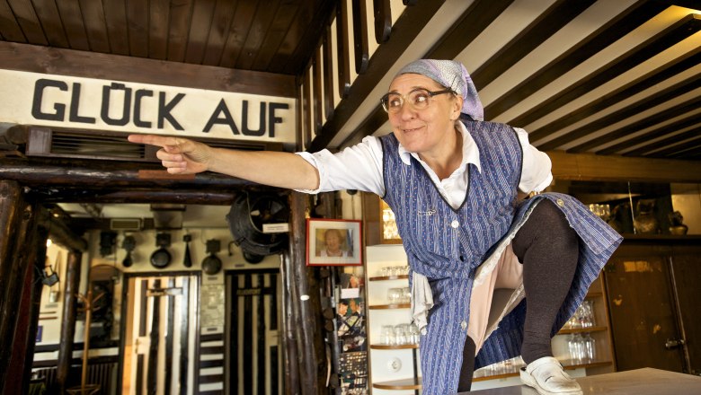 A woman in traditional dress points to something in a rustic room. "Gl&uuml;ck Auf" is written above her.