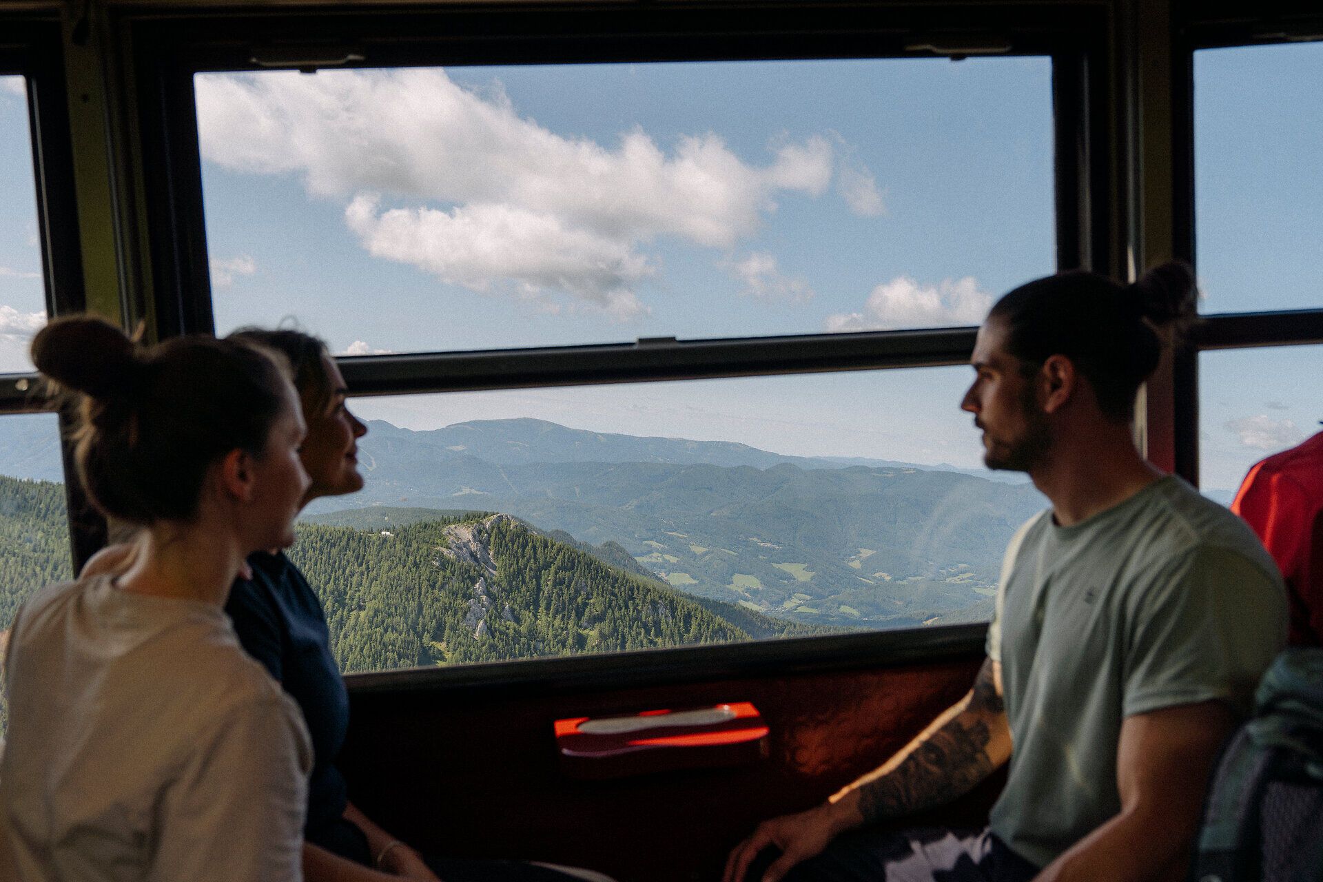 Zwei fröhliche Frauen schauen aus dem Fenster des Salamander-Zuges der Schneebergbahn, während die Landschaft aus Bäumen im Hintergrund vorbeizieht, was Freude und Neugier auf das bevorstehende Reiseerlebnis widerspiegelt.
