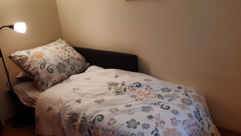 Ein kleines Zimmer mit einem Einzelbett, das mit einer gebl&uuml;mten Bettdecke bezogen ist. Eine Stehlampe steht daneben, und ein buntes Bild h&auml;ngt an der Wand.