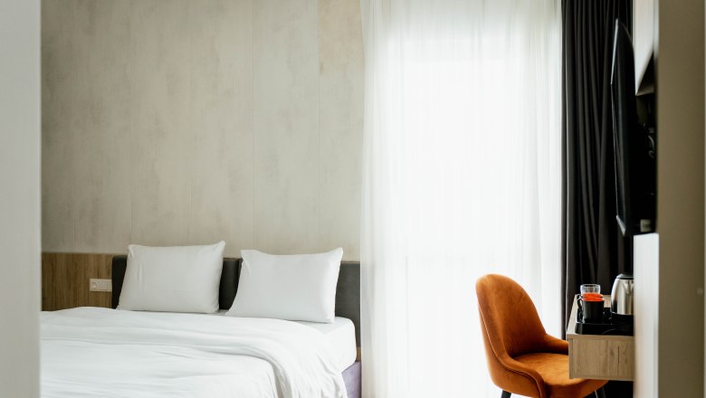 Modernes Hotelzimmer mit Doppelbett, weißen Bettlaken, Holzregal und orangefarbenem Stuhl vor einem Fenster mit Vorhängen.