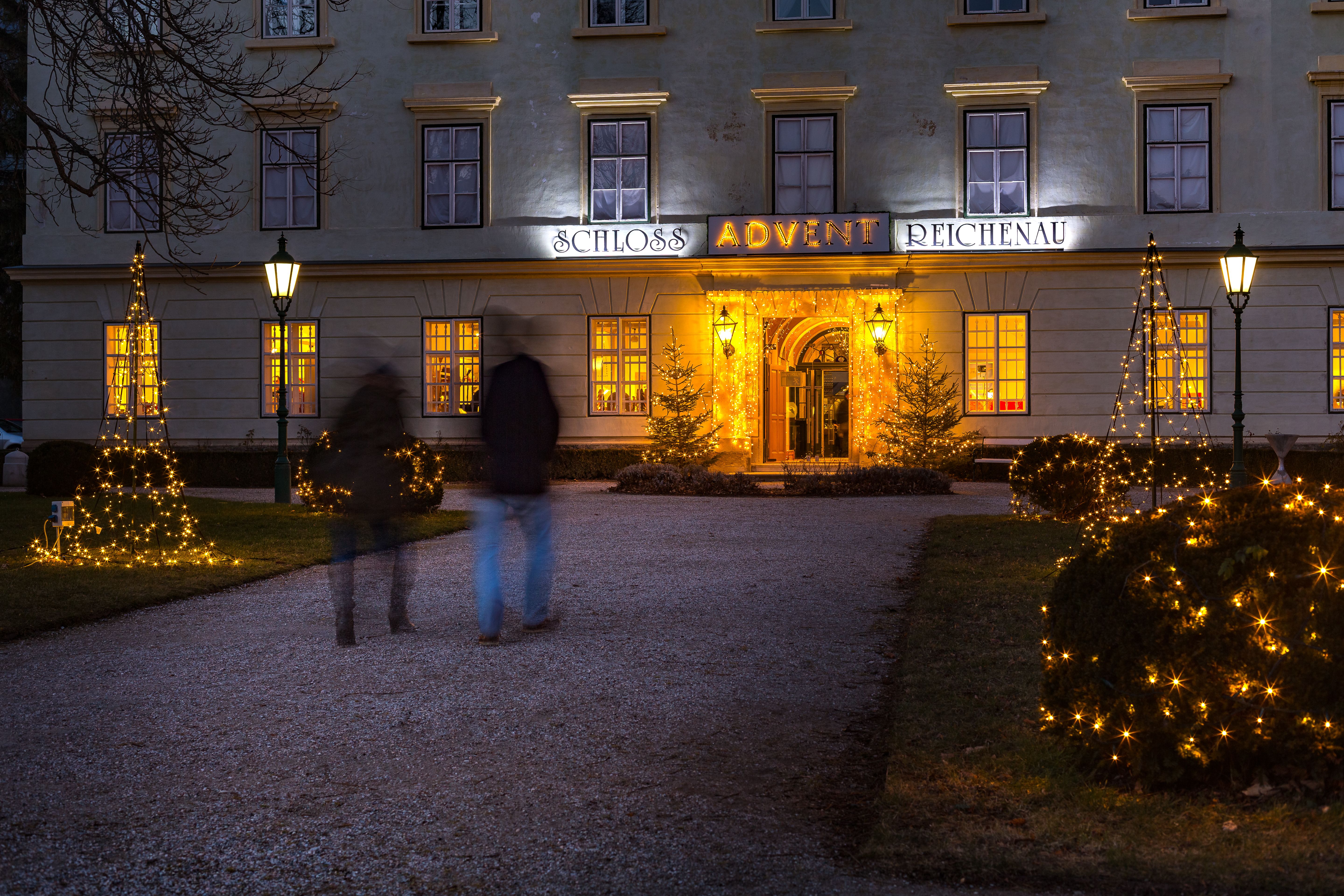 Adventmarkt im Schloss Reichenau an der Rax. Das Schloss ist von außen stimmungsvoll beleuchtet, im Innenhof stehen die Hütten. Menschen stehen davor und trinken Punsch.
