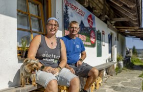 Zwei Personen sitzen vor einem Gebäude mit einem Hund, im Hintergrund ein Plakat mit Datum 09. Sept. 2023.