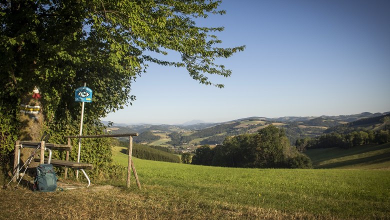Landschaft mit Bank, Wanderstock und Rucksack im Vordergrund, Hügel und Berge im Hintergrund.