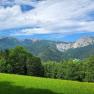 Mountain landscape with green meadows and wooded hills under a blue sky.