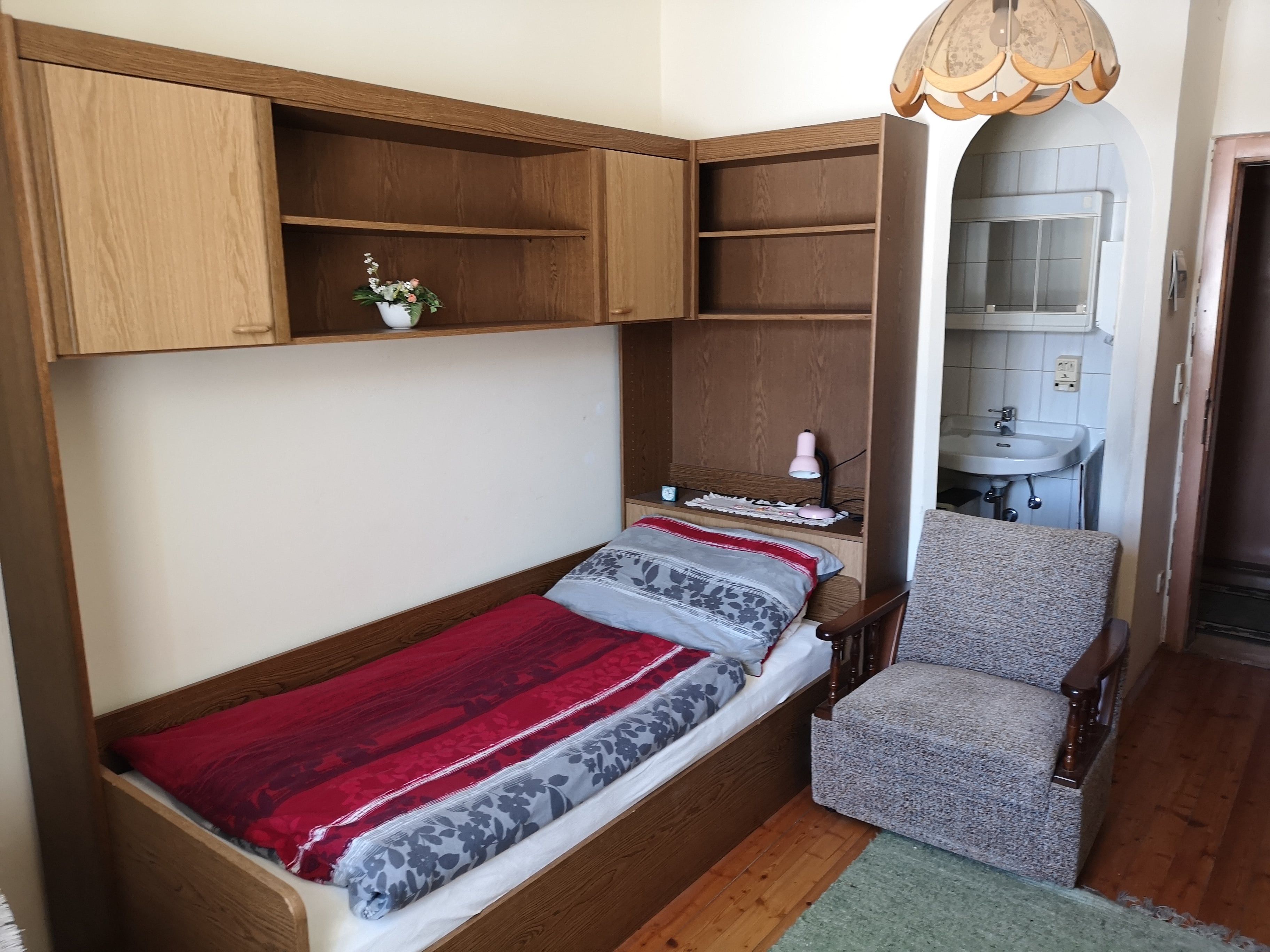 Einzelbett mit roter und grauer Bettwäsche, daneben ein Sessel und ein Waschbecken im Hintergrund.