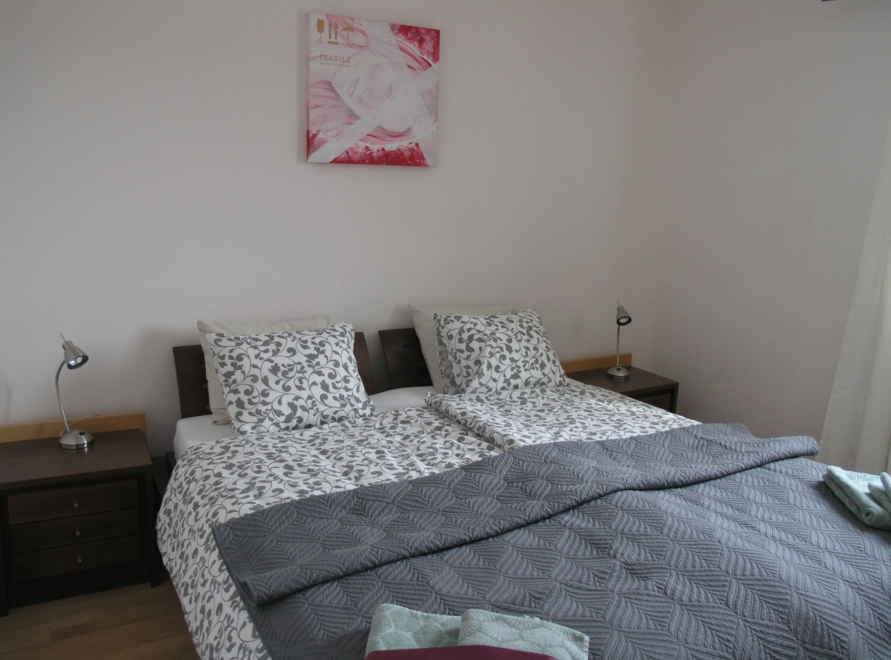 Ein Doppelbett mit gemusterten Kissen und grauer Decke, flankiert von Nachttischen mit Lampen. An der Wand hängt ein Bild.
