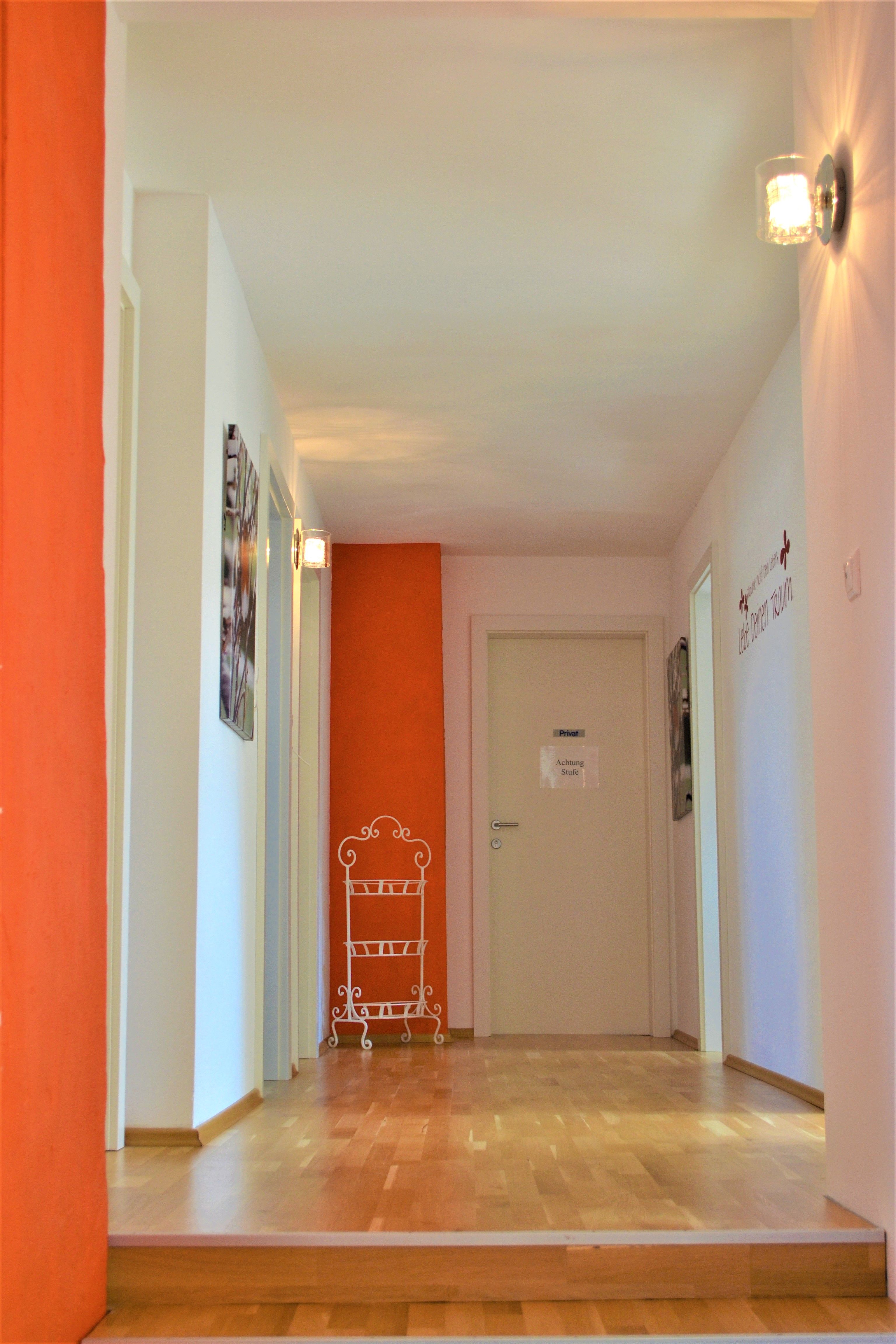 Ein Flur mit Holzboden, weißen Wänden und einer orangefarbenen Wand. An der Wand hängt ein Schild mit der Aufschrift 'Achtung Stufe'.