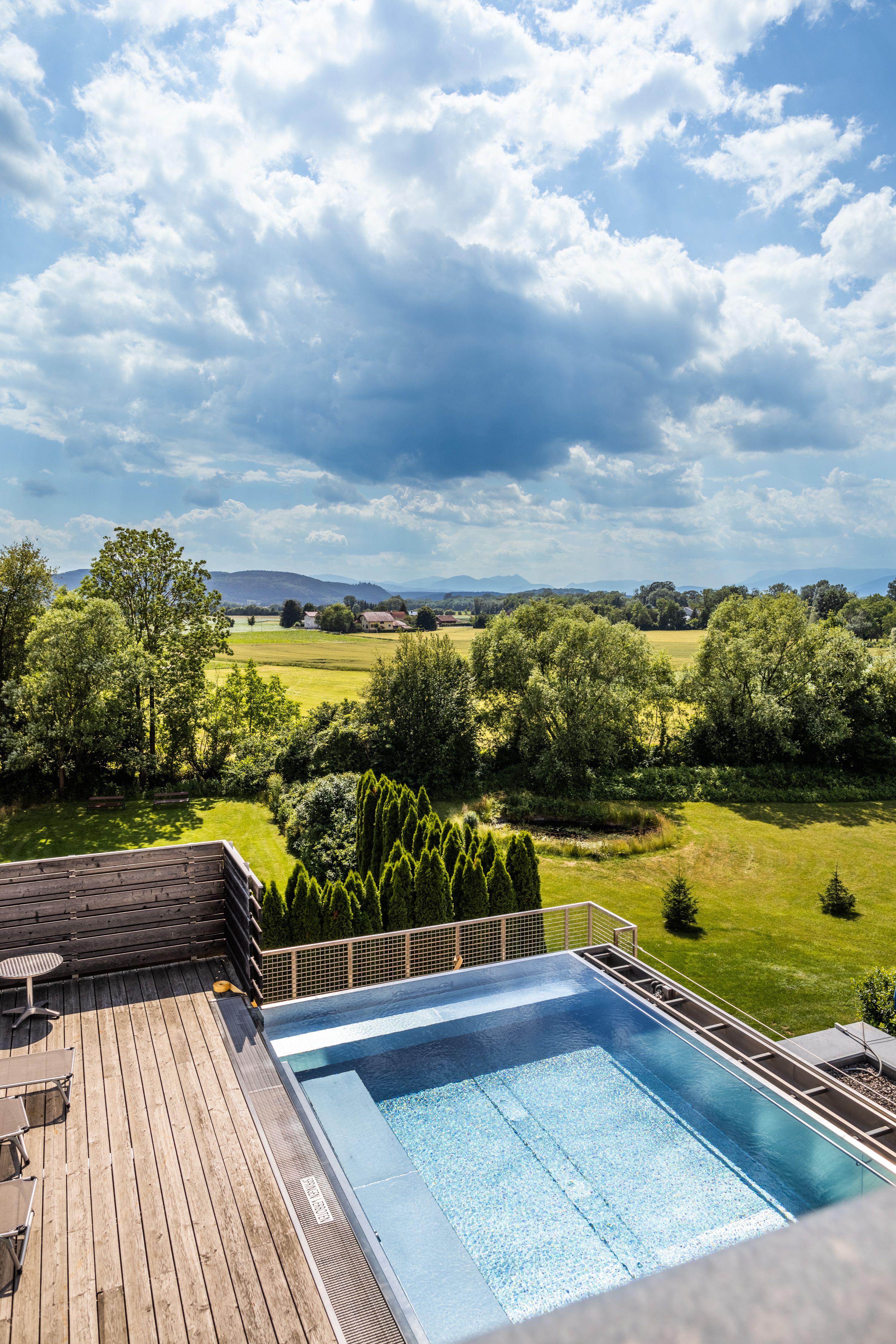 Ein moderner Pool auf einer Terrasse mit Blick auf eine grüne Landschaft und bewölkten Himmel.