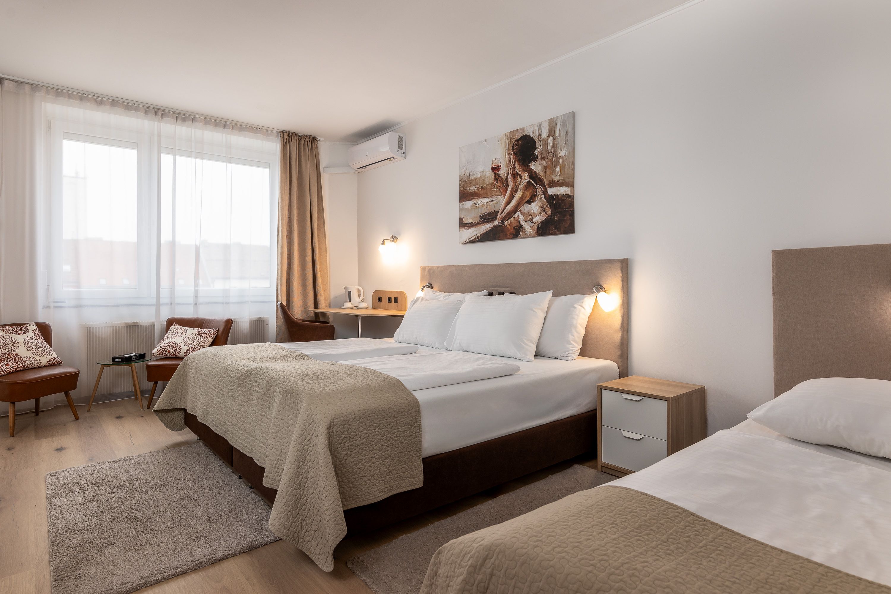 Helles Zimmer mit 2 Doppelbetten, beigen Vorhängen und Teppich, Holzboden, Nachtkästchen und Sitzmöglichkeiten