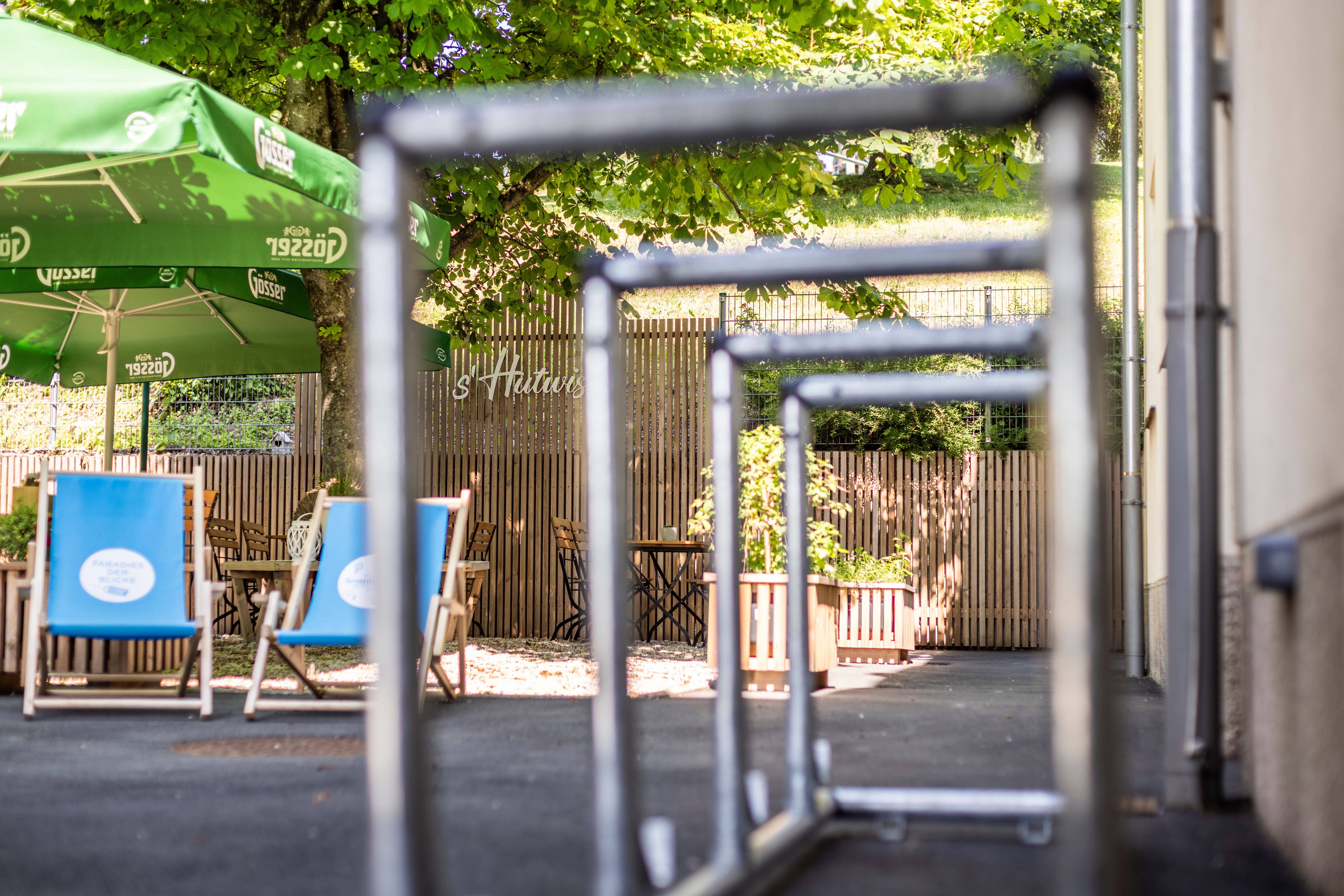 Silberner Fahrradständer, im Hintergrund der Gastgarten im s'Hutwisch mit großen Sonnenschirmen, blauen Liegestühlen, Holzmöbeln und Holzvertäfelung. In der Mitte wächst ein Kastanienbaum.