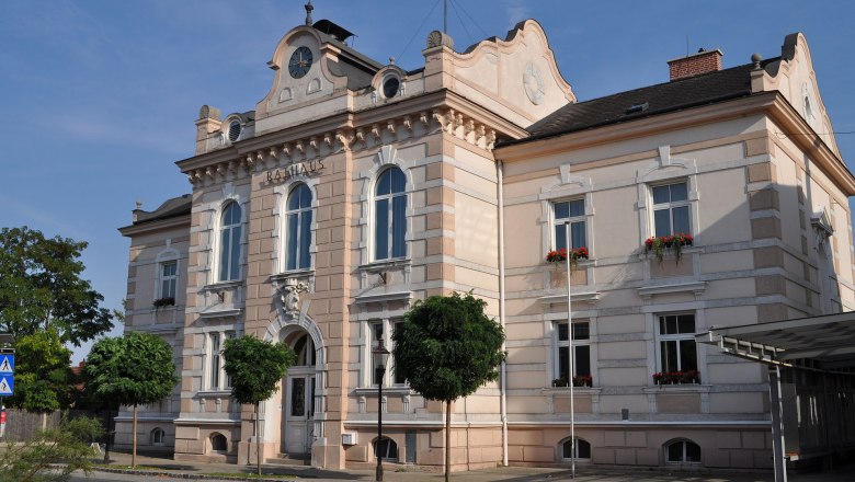 Das Rathaus von Felixdorf, ein historisches Gebäude mit Uhrturm und verzierten Fenstern, umgeben von Bäumen und einer Straße im Vordergrund.