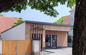 A modern village store with a wooden fa&ccedil;ade and signs, surrounded by trees.