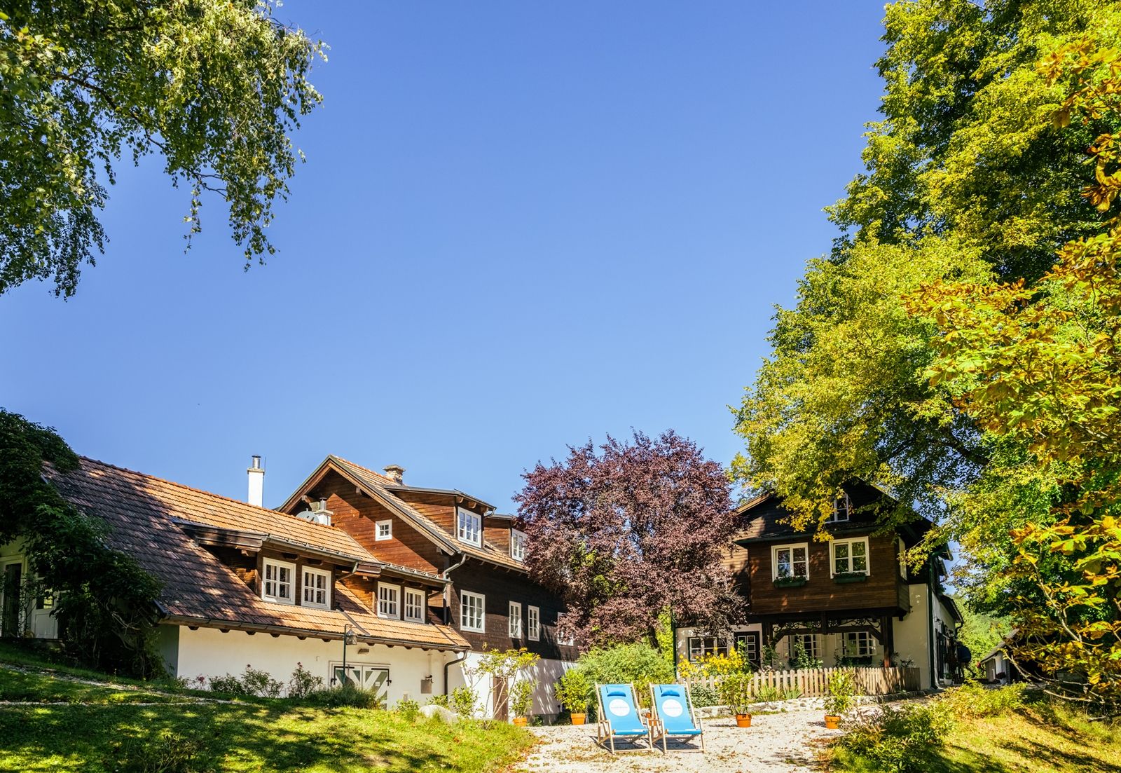Ein traditionelles Landhaus mit Holzdach und blauen Liegestühlen im Garten, umgeben von Bäumen und blauem Himmel.