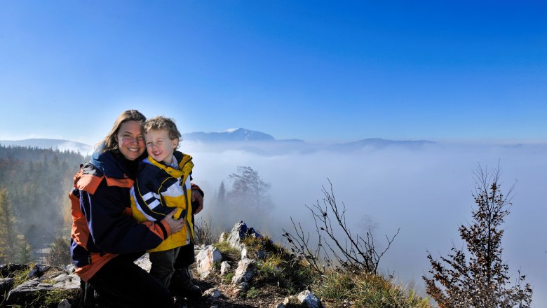 Eine Frau und ein Kind stehen auf einem Hügel mit Blick auf ein Nebelmeer und Berge im Hintergrund.