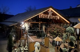 Ein dekorativ beleuchtetes Holzgebäude mit der Aufschrift 'Deko Stadl' bei Nacht, umgeben von Pflanzen und Dekorationsartikeln.