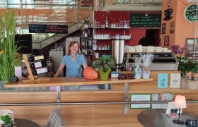 Eine Frau steht hinter der Theke eines Cafés mit Pflanzen, Weinflaschen und einer Kaffeemaschine.
