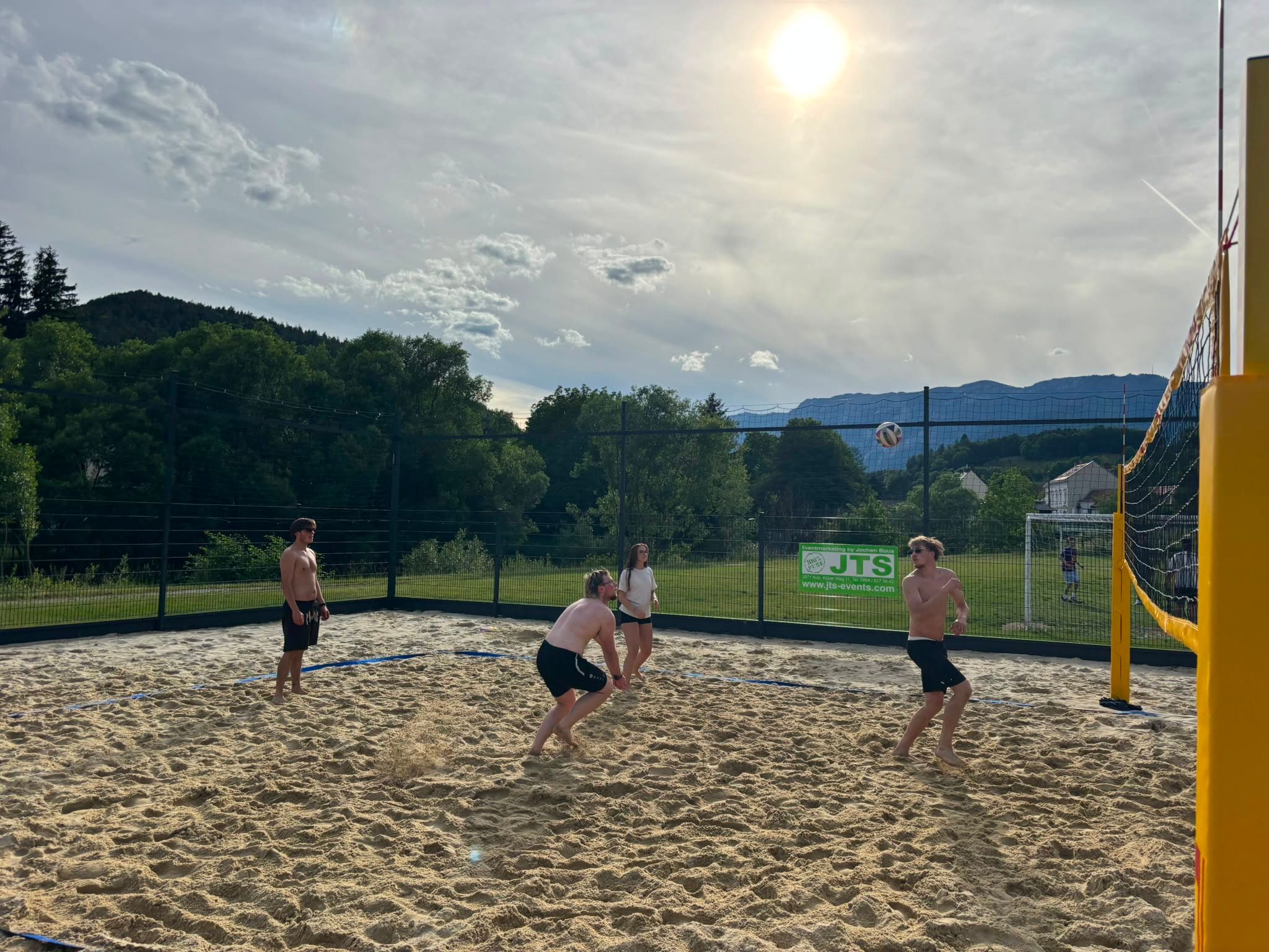 Four people play beach volleyball on a sandy court in sunny weather, surrounded by trees and mountains.