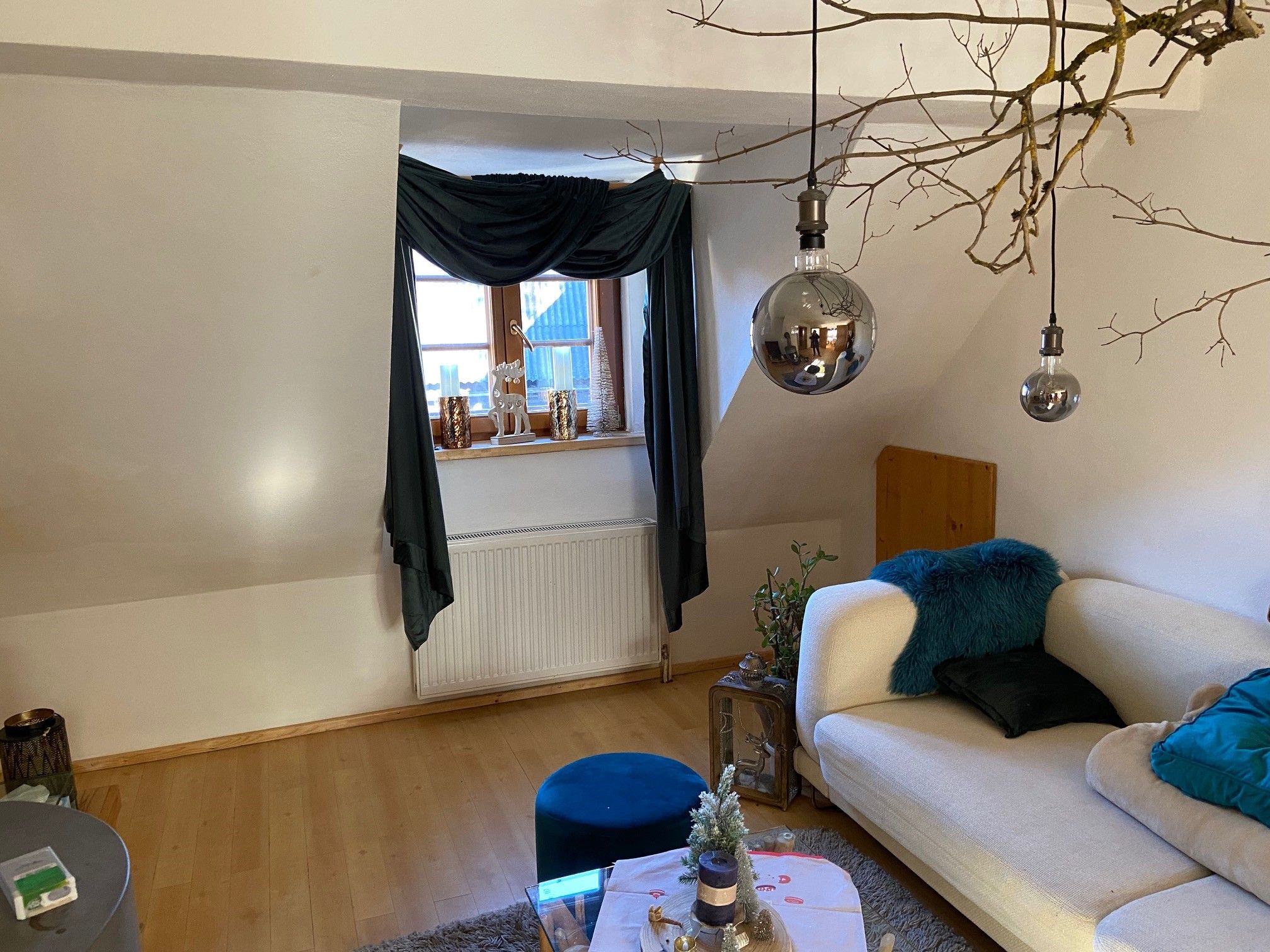 Gemütliches Wohnzimmer mit Sofa, Holzboden, Fenster mit Vorhängen, dekorativen Kerzen und hängenden Lampen.