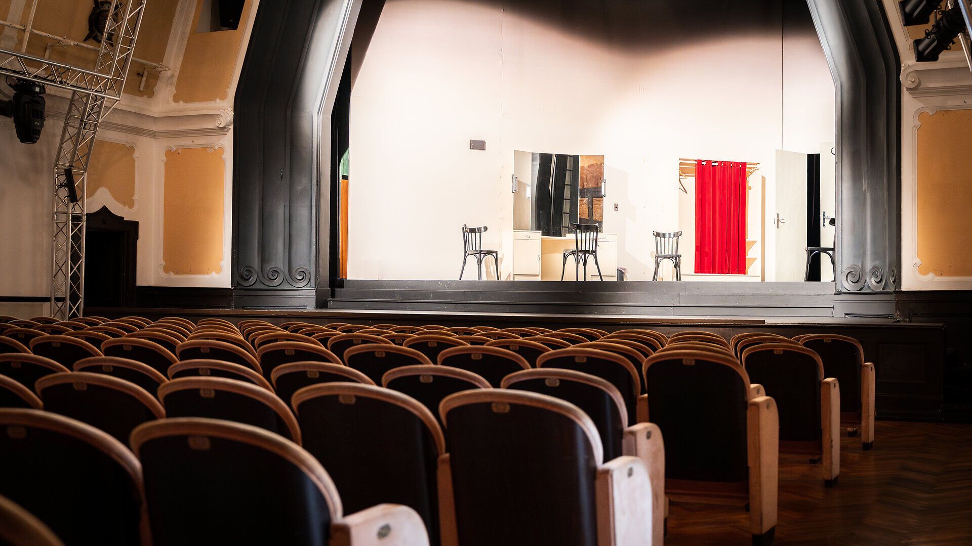 Theatersaal mit BLick zur Bühne, auf der Sessel, ein Spiegel und ein roter Vorhang stehen.