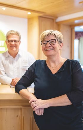 Petra Weber steht vor der Rezeption aus Holz im VitalZeit Hotel Weber, hinter der Rezeption steht Alfred Weber. Im Hintergrund hängt das grüne, runde Logo des Hotels und man sieht Schlüssel.