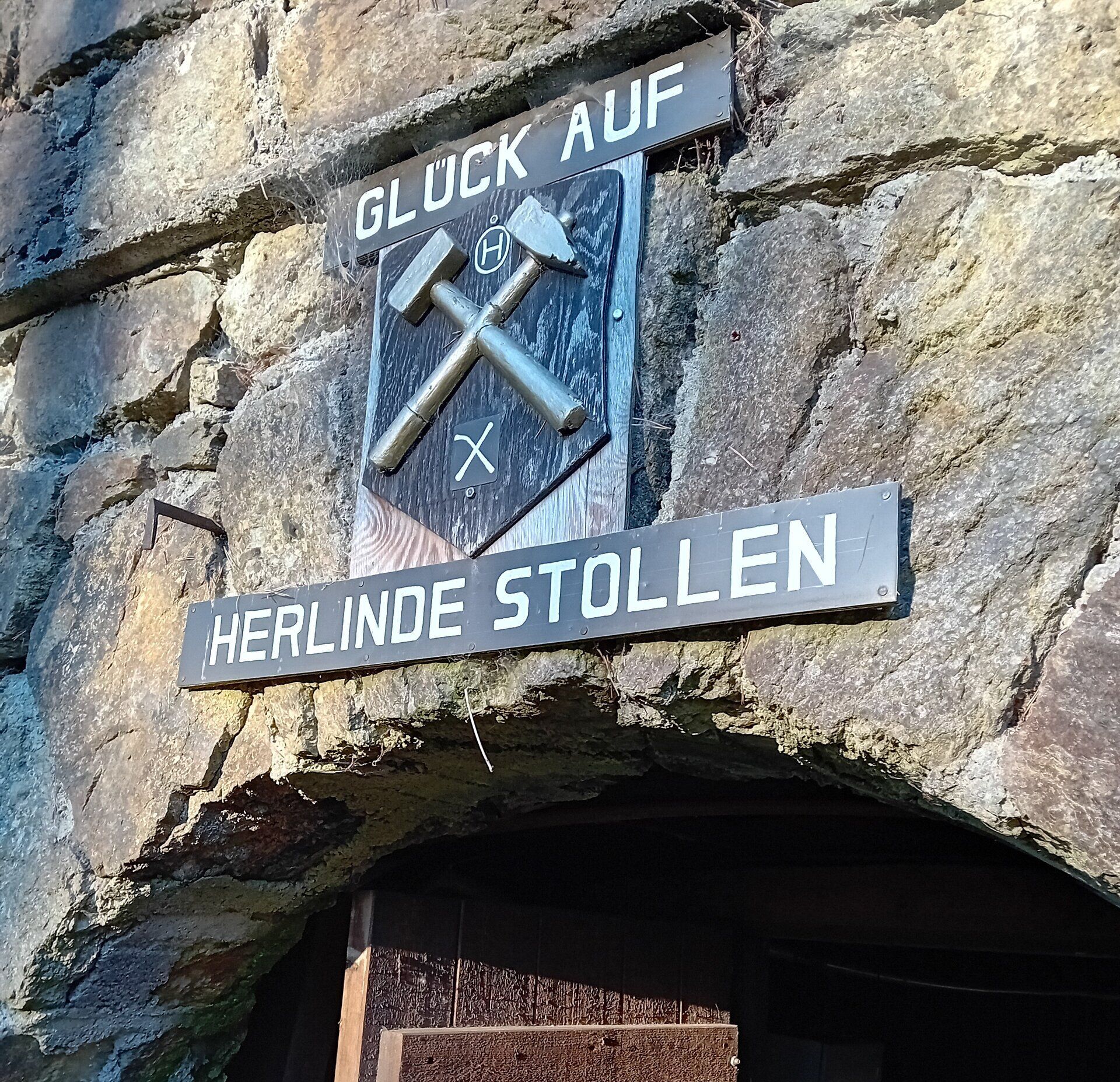Eingang und Innenansicht des historischen Bergwerks „Herlinde Stollen“ mit Schienen und Grubenschild „Glück auf“.