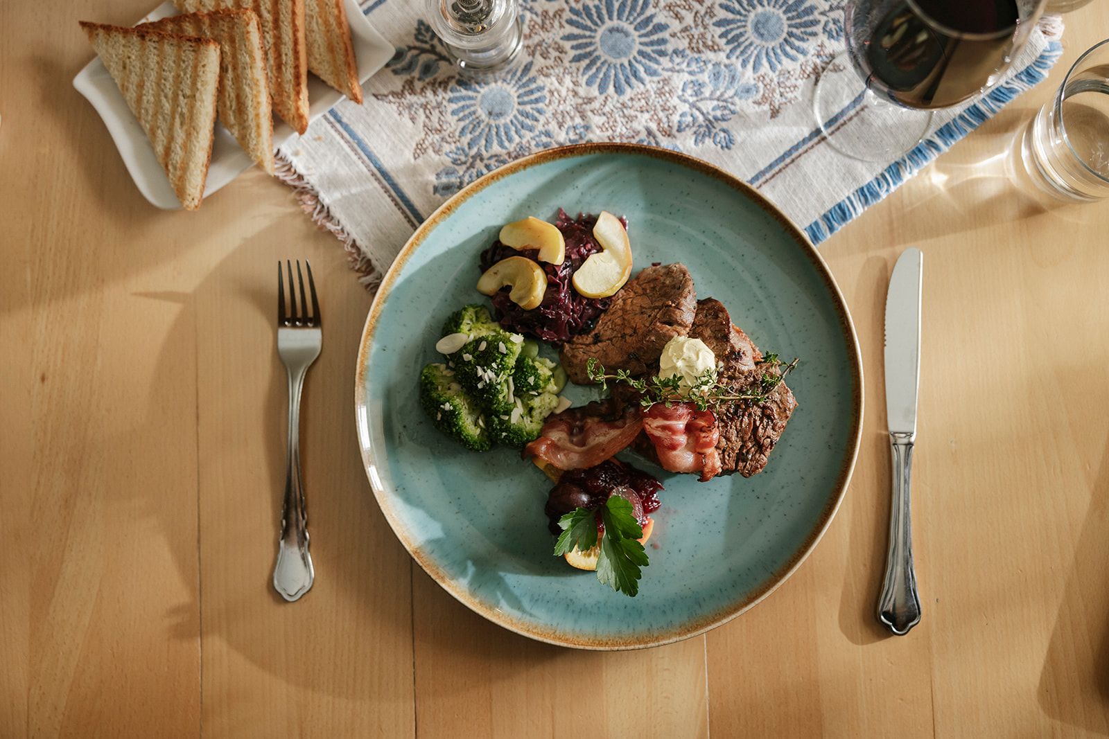angerichtete Speise mit Fleisch, Speck, Rotkraut und Brokkoli auf einem blauen Teller, daneben gedecktes Besteck