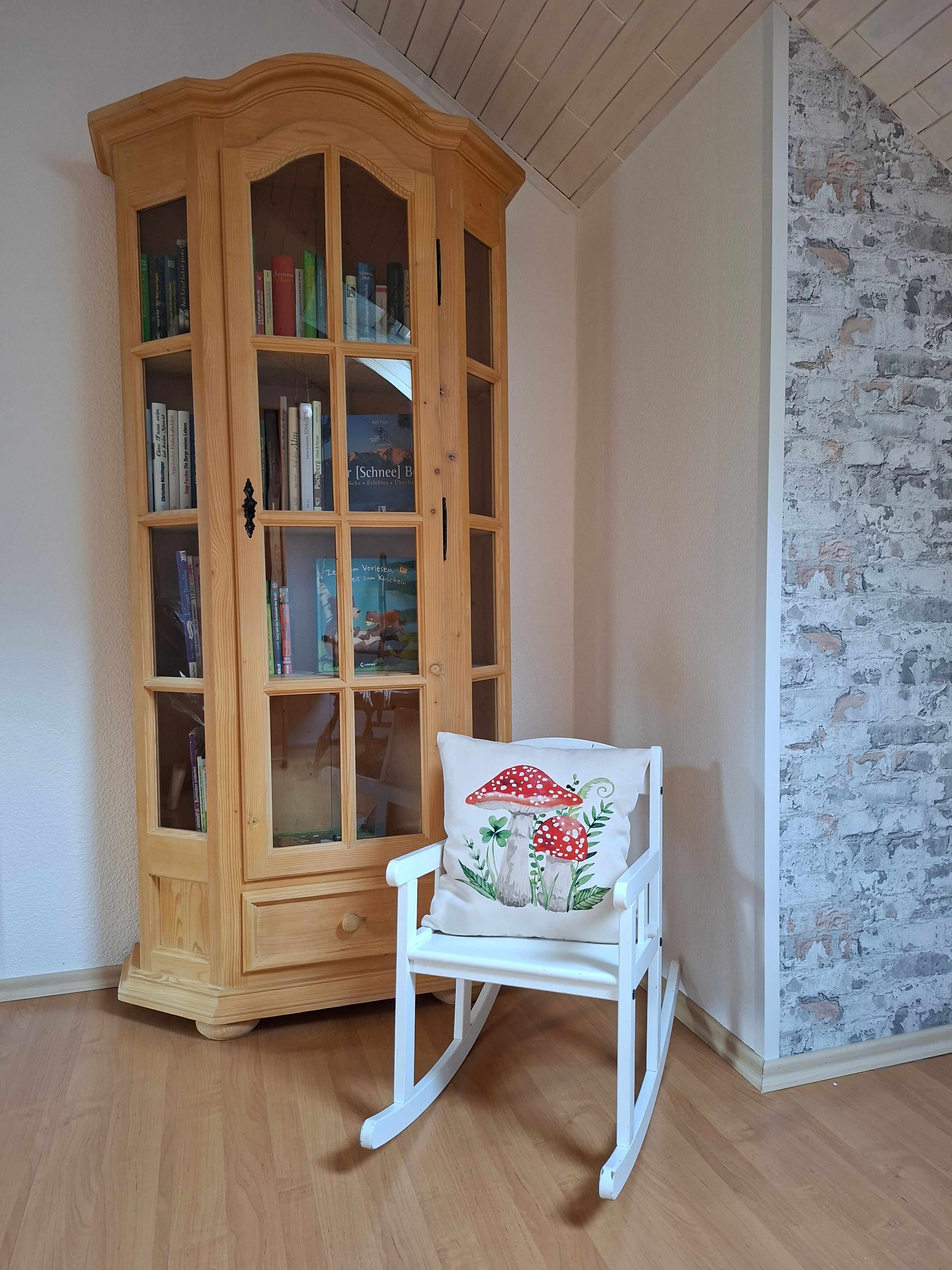 Ein Holzschrank mit Büchern und ein weißer Schaukelstuhl mit einem Kissen, das Pilze zeigt, stehen in einem Raum mit Holzboden und gemusterter Tapete.