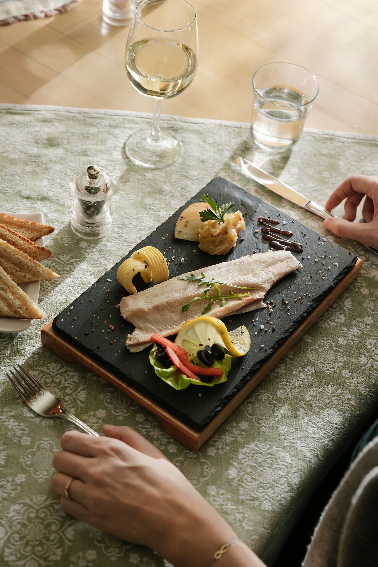 Angerichteter Teller mit geräuchertem Fisch mit Garnierung, dahinter steht ein Glas Wein und auf einem Teller liegen getoastete Brotscheiben.