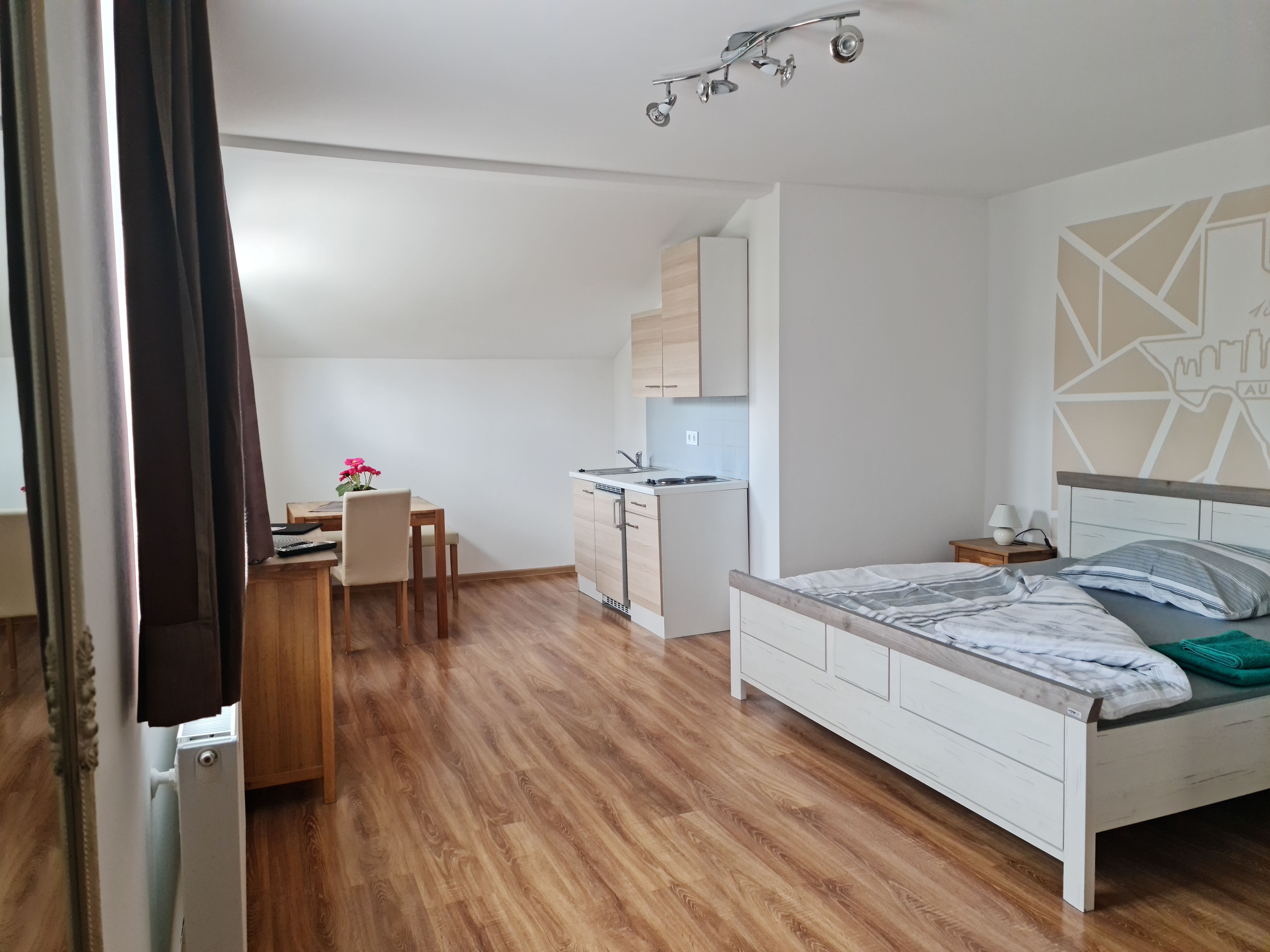 Helles Zimmer mit Holzboden, Bett, Esstisch und kleiner Küchenzeile.