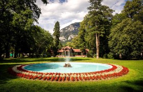 Inmitten einer malerischen Landschaft sprudelt der Brunnen in einem liebevoll gestalteten Garten, umgeben von bunten Blumenbeeten. Die majest&auml;tischen Berge im Hintergrund verleihen der Szenerie eine beeindruckende Kulisse, die zum Verweilen und Entspannen einl&auml;dt.