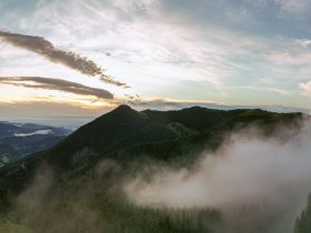 Panoramablick auf den Sonnwendstein bei Sonnenaufgang mit Nebel und einem Aussichtsturm im Vordergrund.